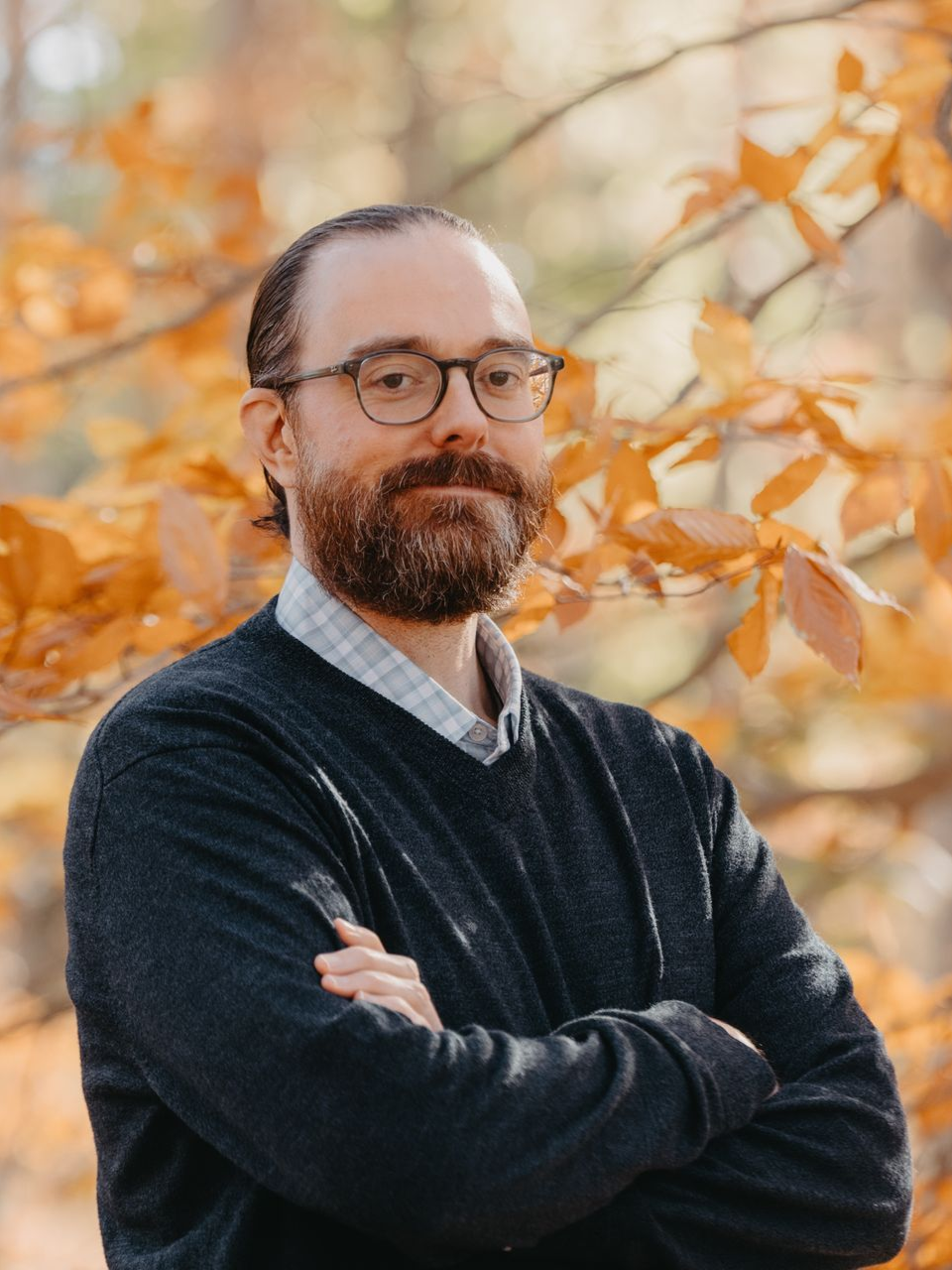 Man with glasses and beard, smiling, against fall foliage backdrop.
