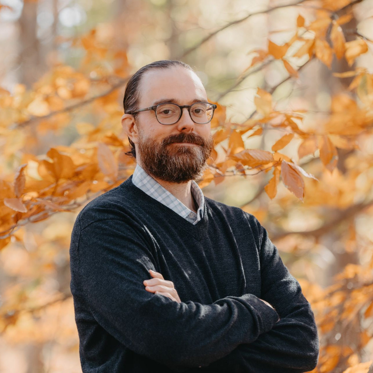 Man with glasses and beard, arms crossed, stands in front of fall foliage.