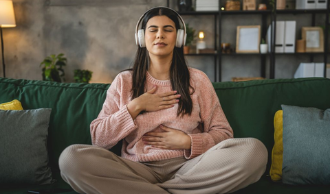 Woman meditating on a green couch, wearing headphones and sweater, eyes closed, hands on chest and stomach.