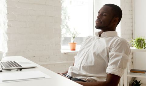 Man sitting at desk, eyes closed, resting. White shirt, laptop, and window in background.