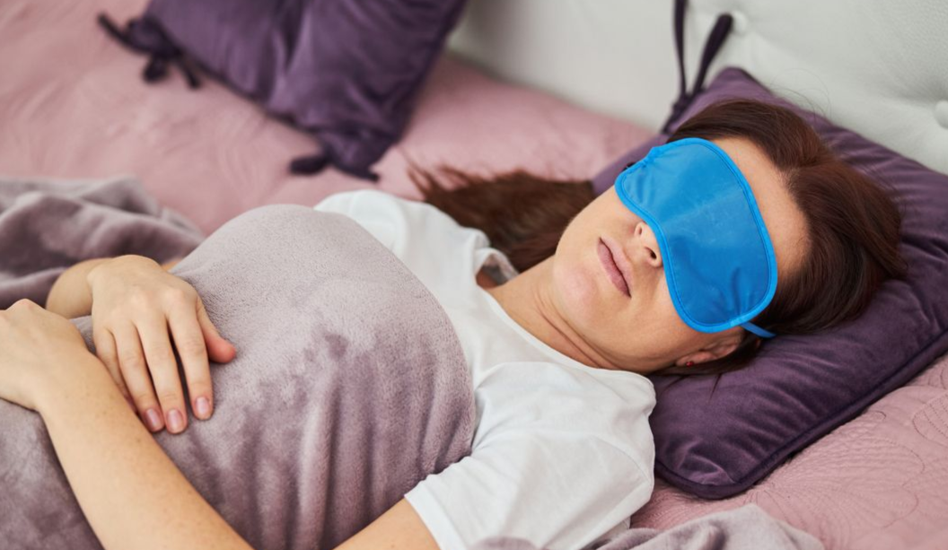 Woman asleep in bed, wearing a blue sleep mask, under a blanket, with purple pillows.