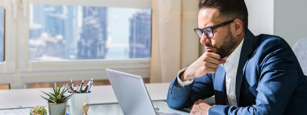 A man wearing glasses looks at a laptop while sitting at a desk with a window in the background.