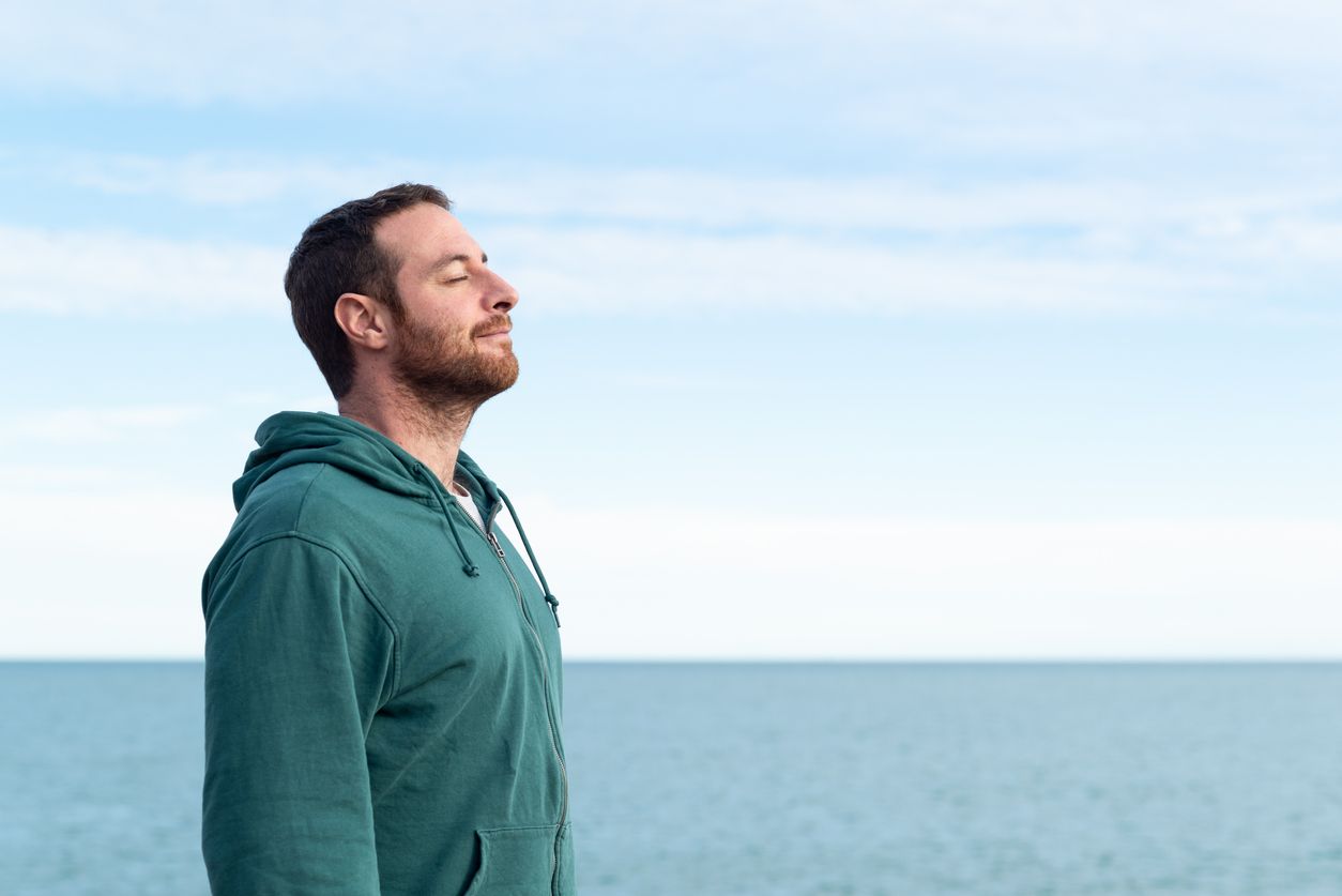Man with closed eyes, outdoors, breaths deeply, blue sky and water in the background.