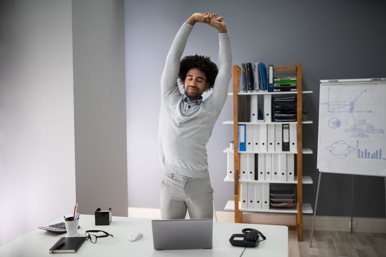 Man stretching at a desk in an office. He has arms above his head and is smiling.