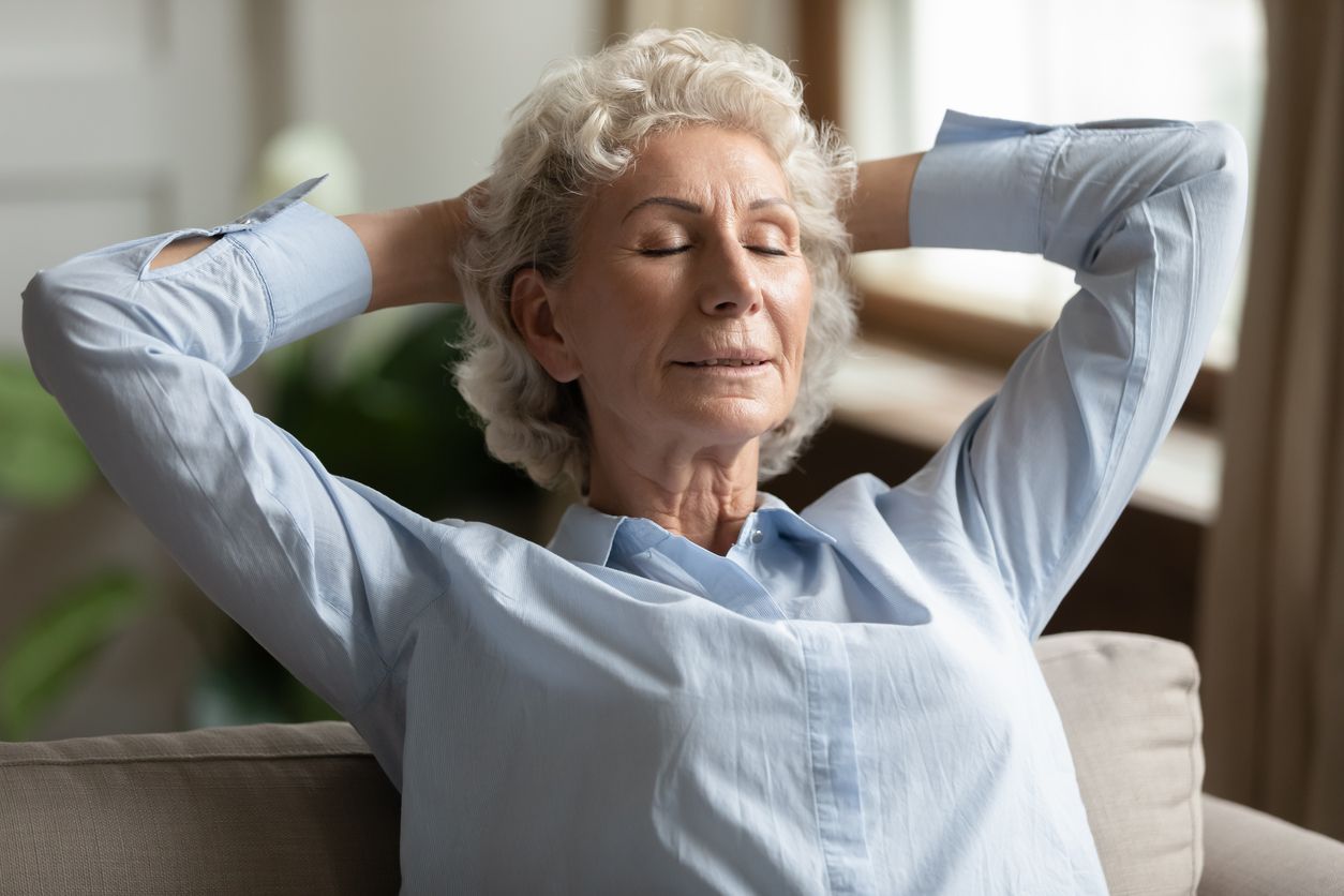 Woman with grey hair, eyes closed, hands behind her head, relaxing on a sofa.
