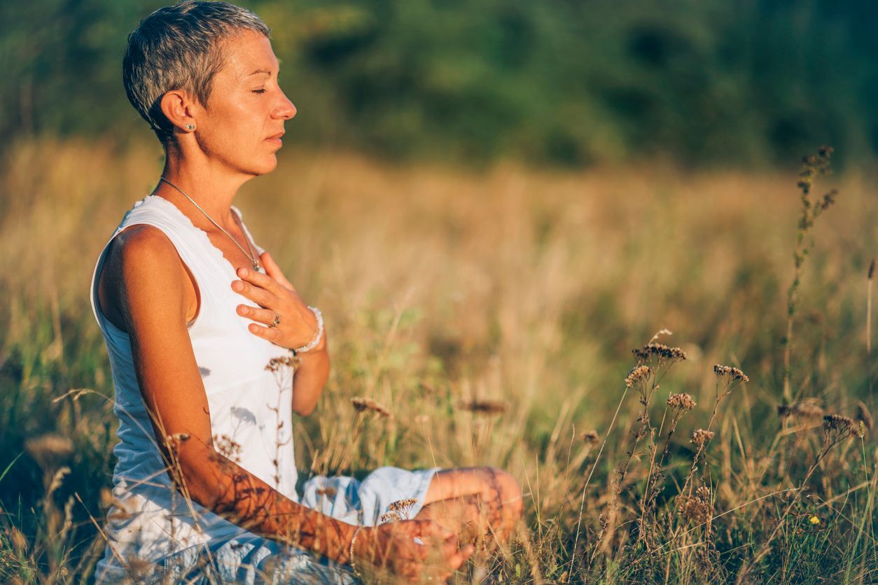 Woman meditating in tall grass, hands on chest, eyes closed, sunny outdoor setting.