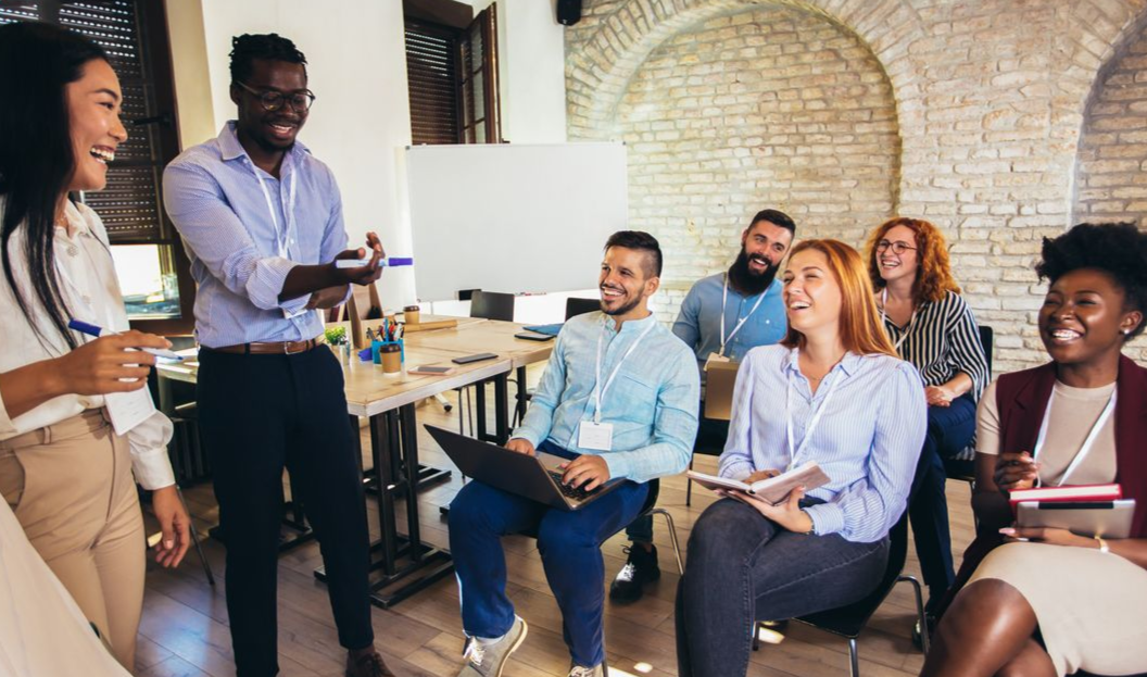 Group of people laughing in a bright office. A man is speaking, gesturing with hands. Others have name tags.