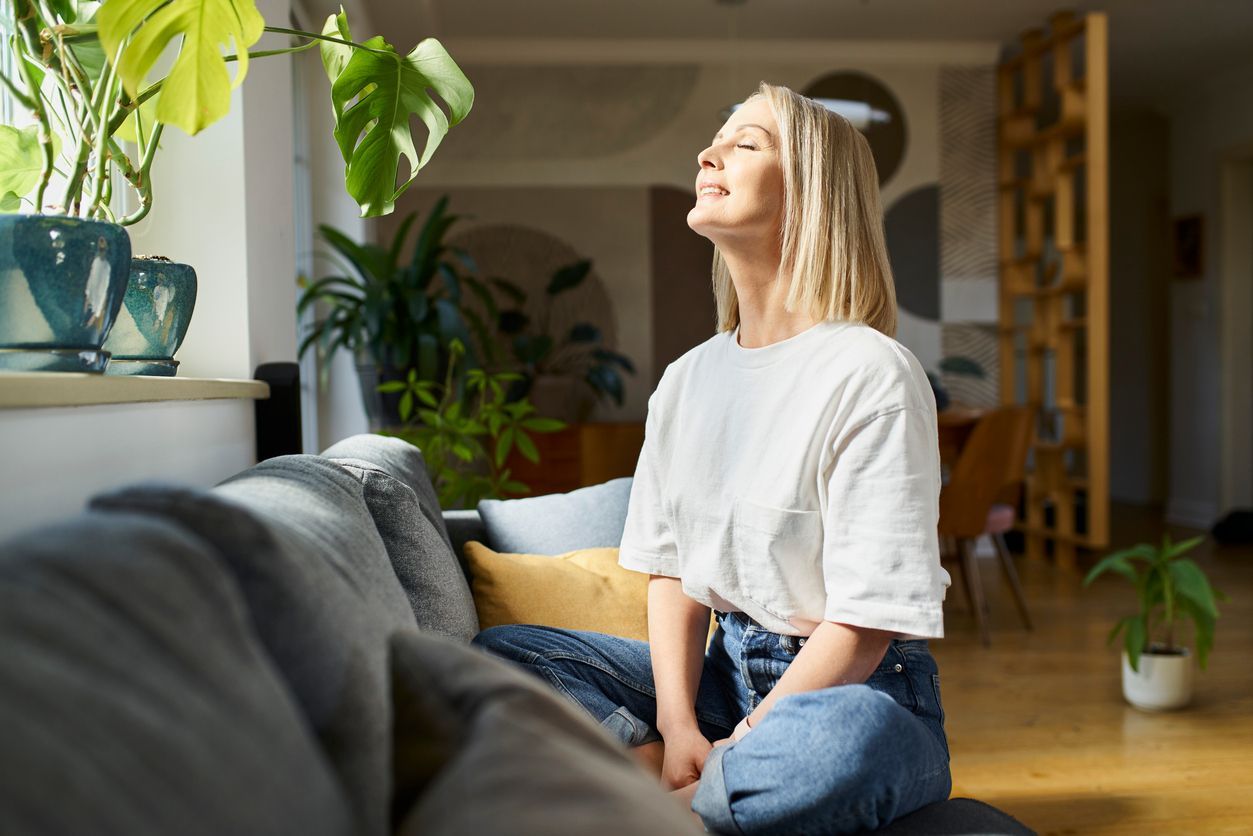 Woman enjoying sunlight on a sofa with indoor plants; smiling.