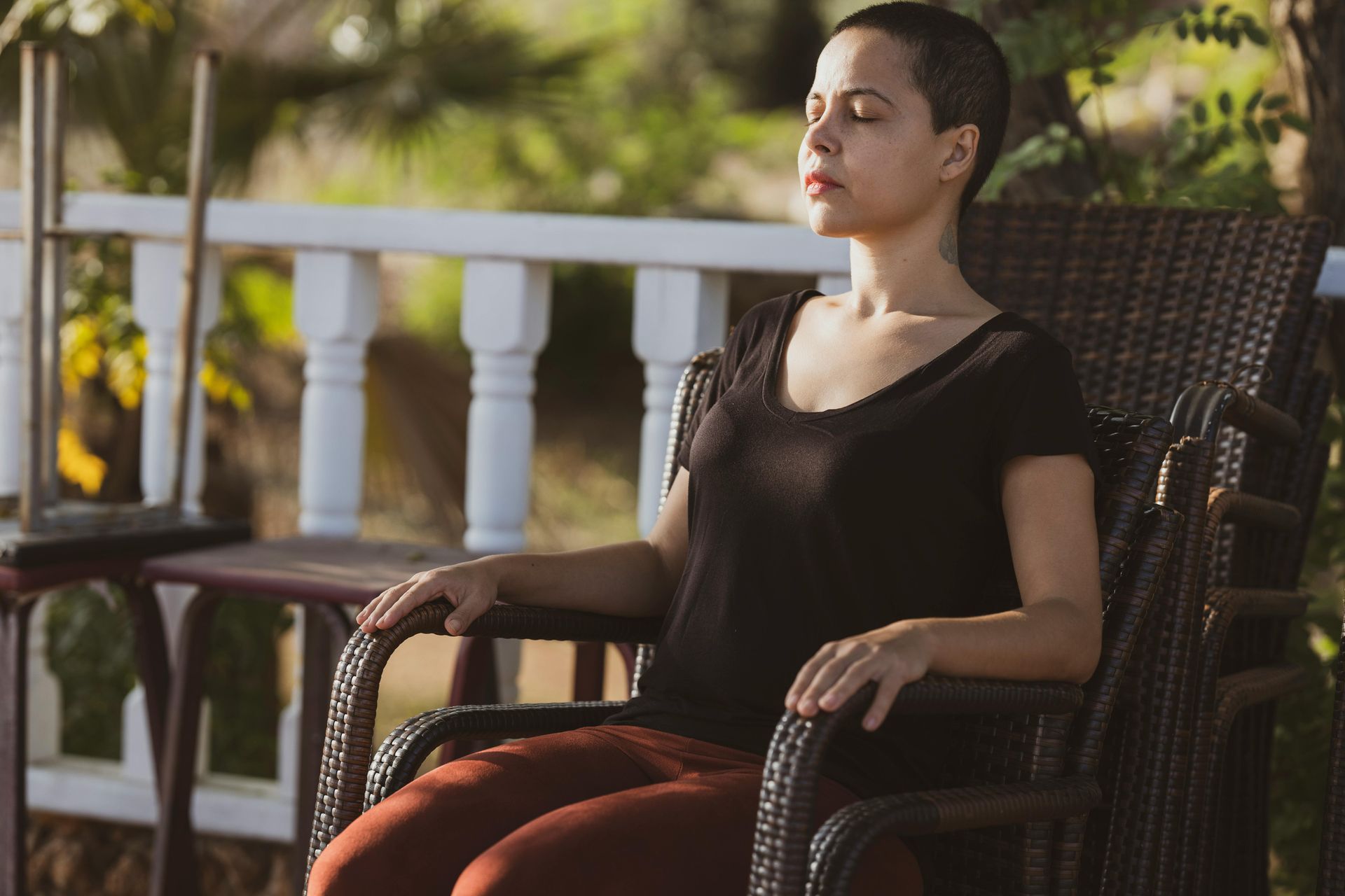 Woman with short hair sitting in a wicker chair, eyes closed, on a porch. Sunny outdoor setting.