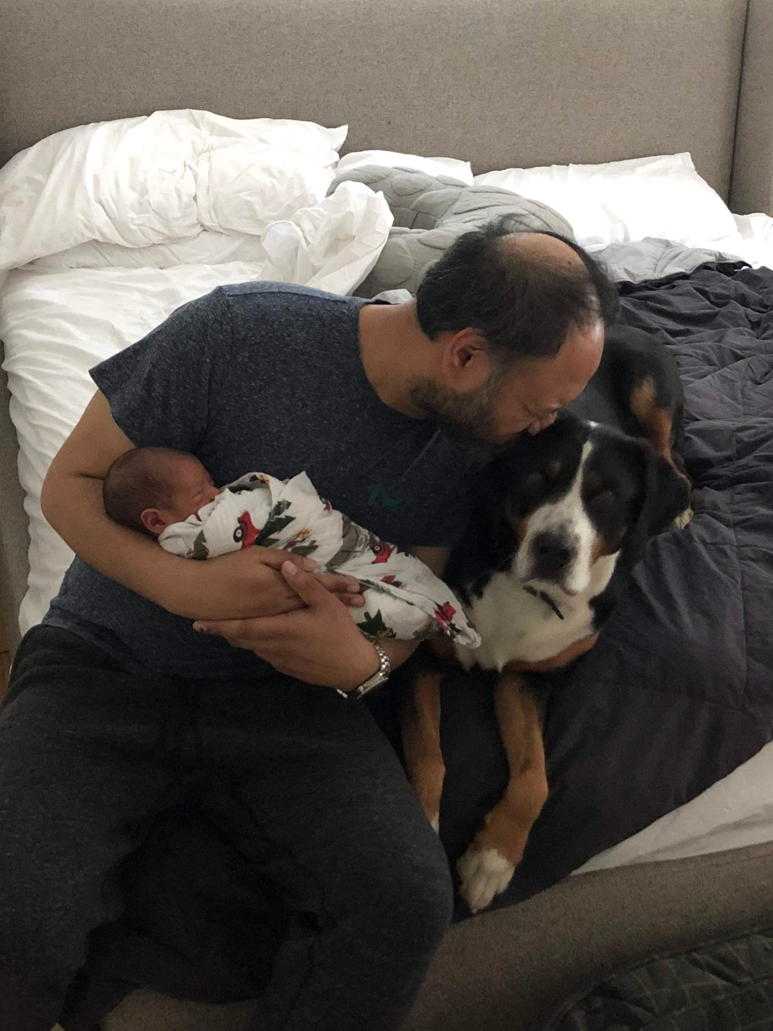A person sitting on a bed holding a swaddled infant while kissing a tricolor dog sitting beside them.