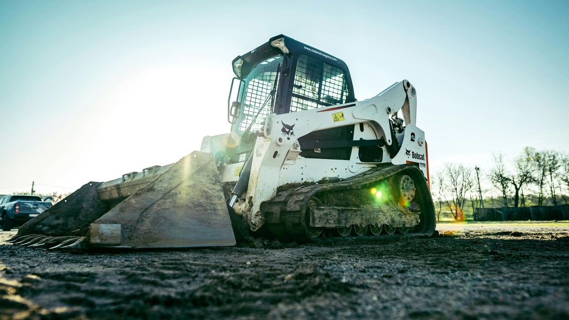A white Bobcat track loader with a full bucket of dirt on a worksite in bright sunlight.