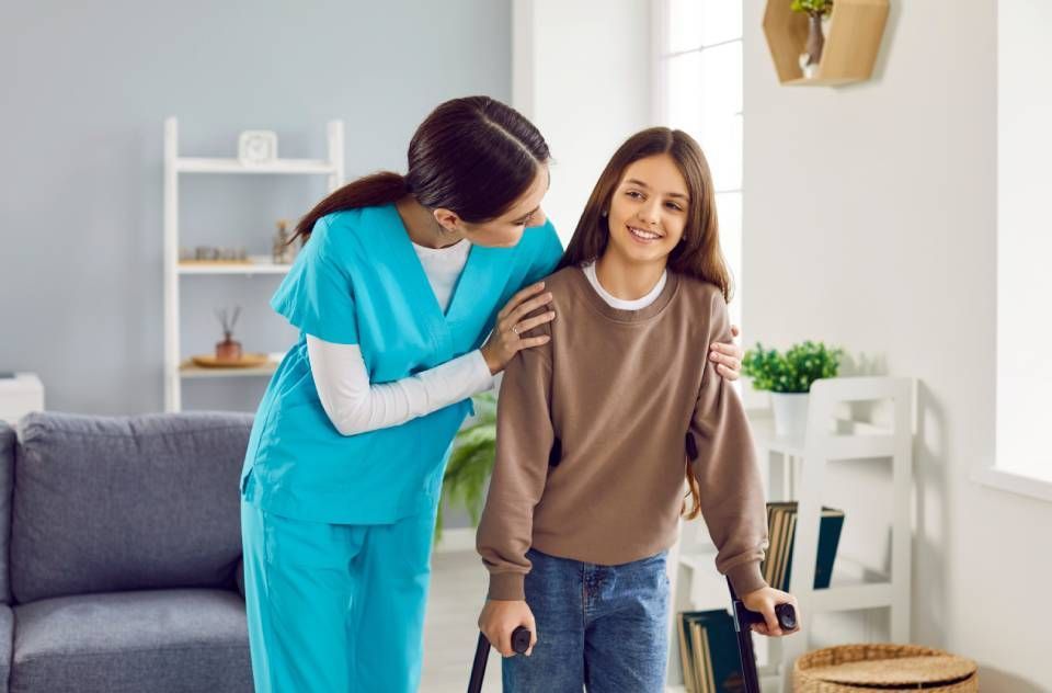 A medical professional assists a smiling person using crutches indoors, blue scrubs, brown sweater.