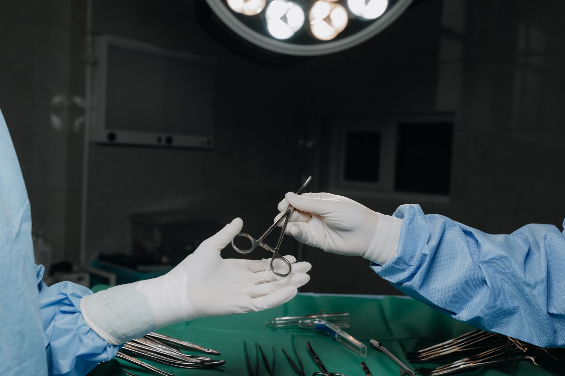 Surgeons' hands in gloves passing surgical tool in a brightly lit operating room.