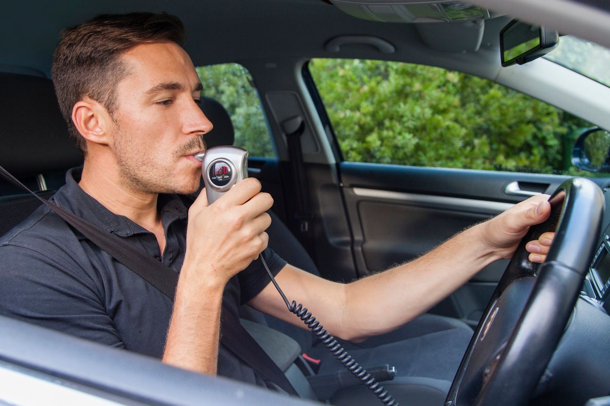 A man is sitting in a car using a breathalyzer.