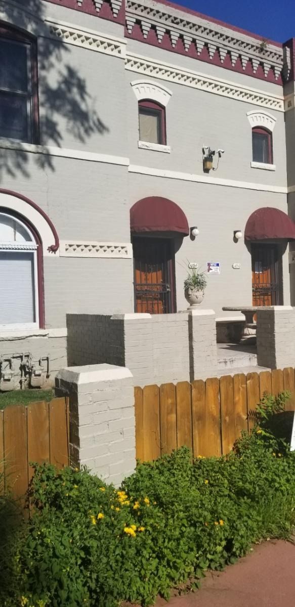 A light gray brick building with dark red awnings, a wooden fence, and green foliage.