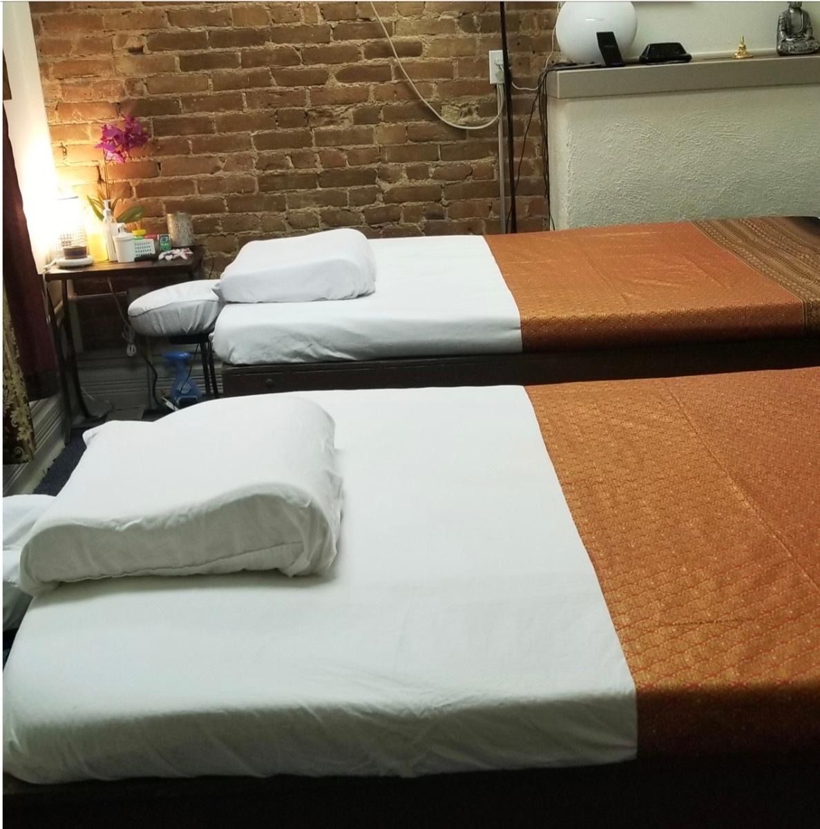 Two massage tables in a room with a brick wall. White sheets, orange covers, and a small side table are visible.