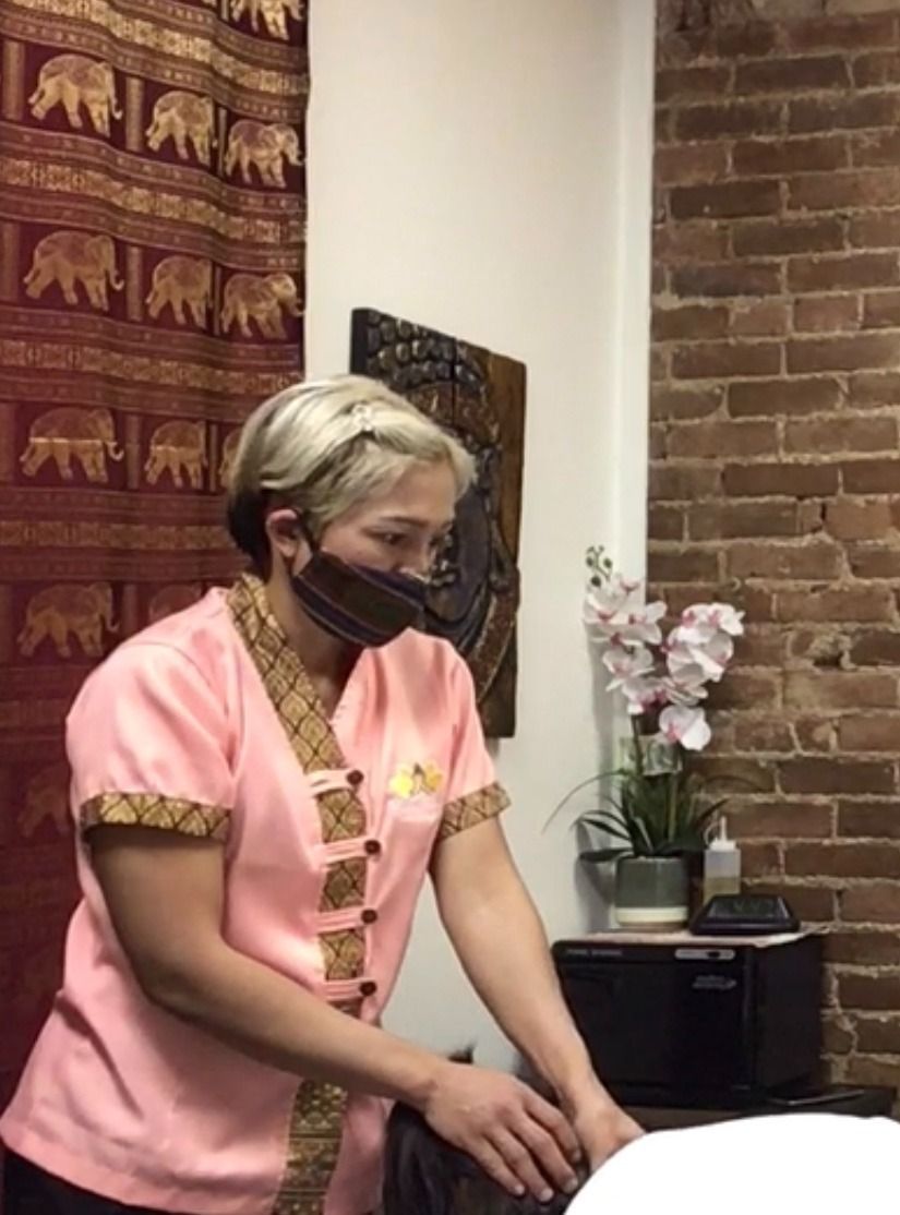 Person in pink massage therapist uniform and face mask, standing near a massage table, in a massage room.