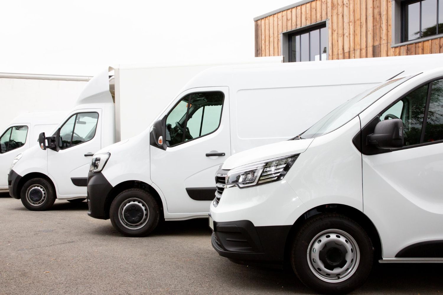 A row of three parked white delivery vans angled in front of a modern building with wood paneling.