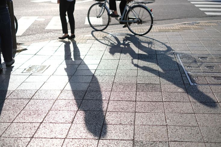 A person stands on a paved sidewalk next to another person riding a bicycle, with their long shadows cast on the ground.