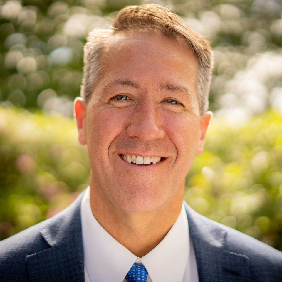 Headshot of a smiling person wearing a navy blue suit, white collared shirt, and blue tie against a soft green background.