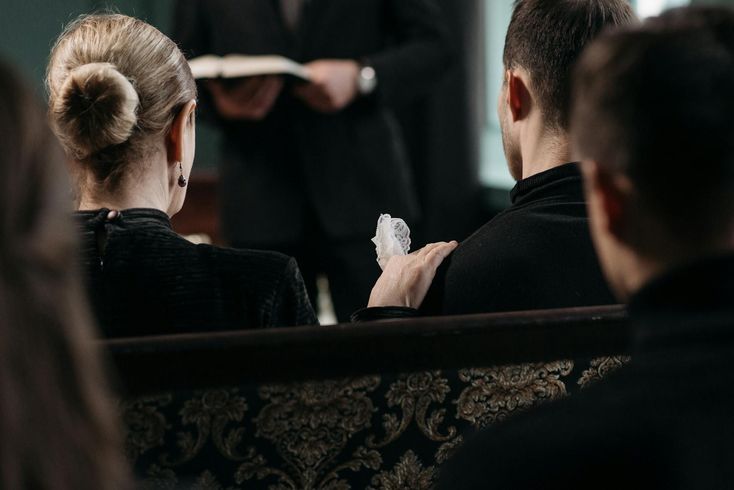 Attendees in dark clothing sit in church pews during a service, with one person resting a hand on another's shoulder.