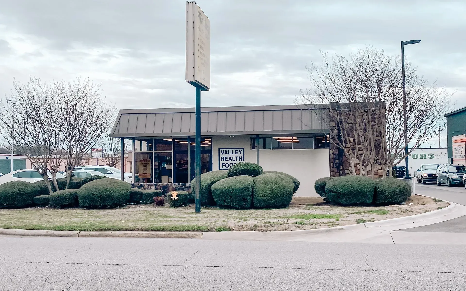Small, light-colored building with a dark roof and a sign, surrounded by shrubs and bare trees on a cloudy day.