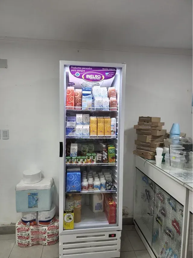 Una pequeña nevera expositora de aperitivos se encuentra en un cuarto de servicio, junto a un dispensador de agua y cajas apiladas.