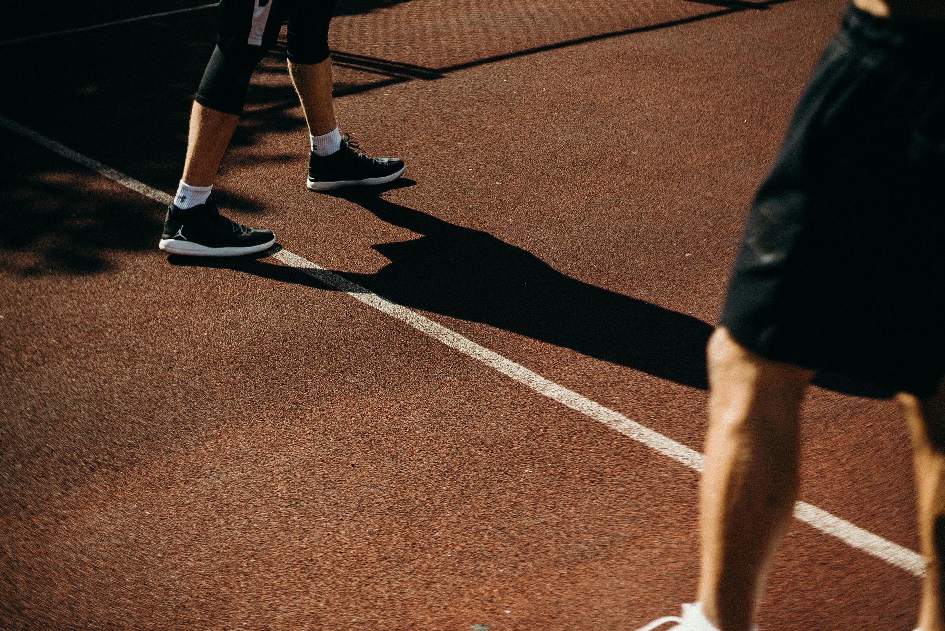 Two people on a red sports court, legs visible, one in black shorts, other in black high-top shoes.