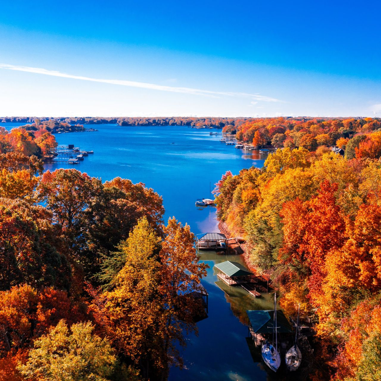Aerial view of a lake surrounded by trees with autumn foliage in shades of orange, yellow, and red.