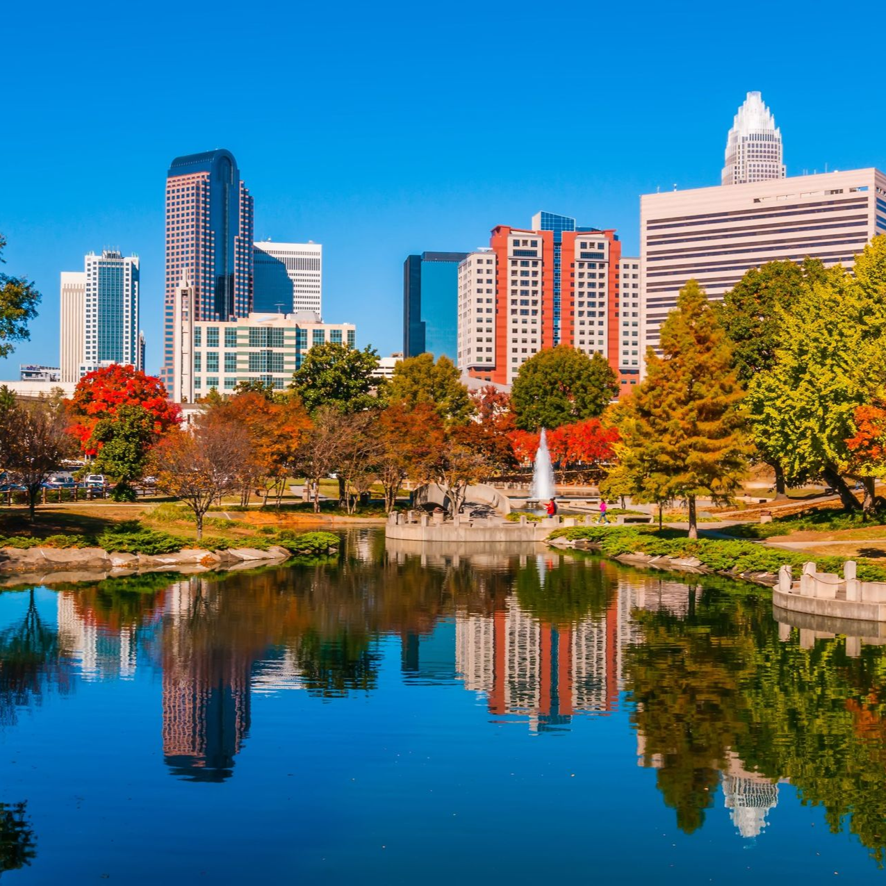 Charlotte, North Carolina skyline reflected in a lake with colorful autumn trees under a clear blue sky.