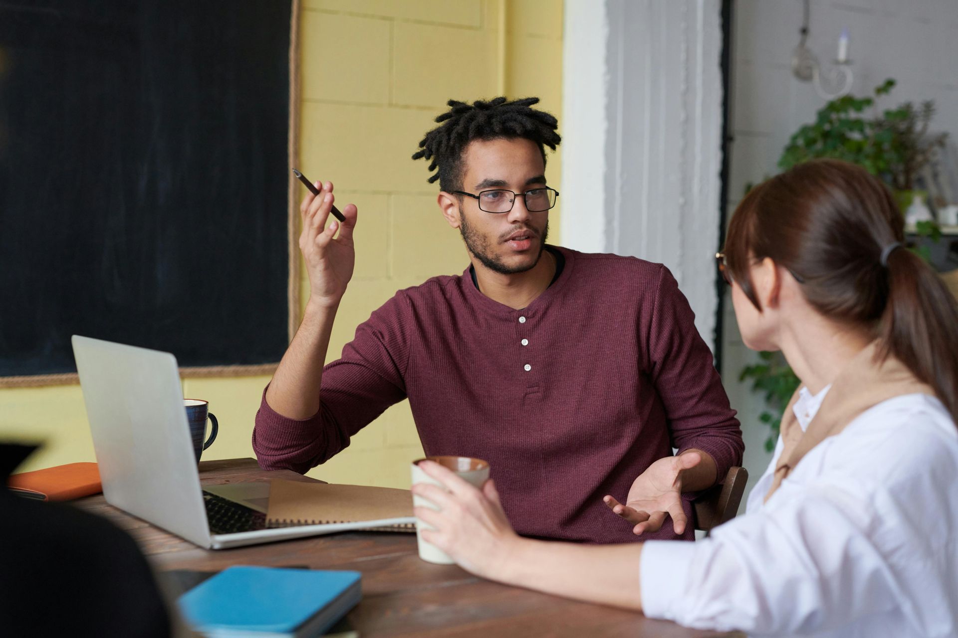 Man gesturing while speaking to a woman at a table, laptop and mug present.