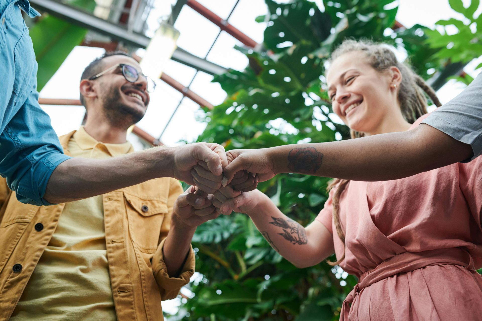People fist bumping, smiling joyfully in a greenhouse setting, surrounded by lush greenery.