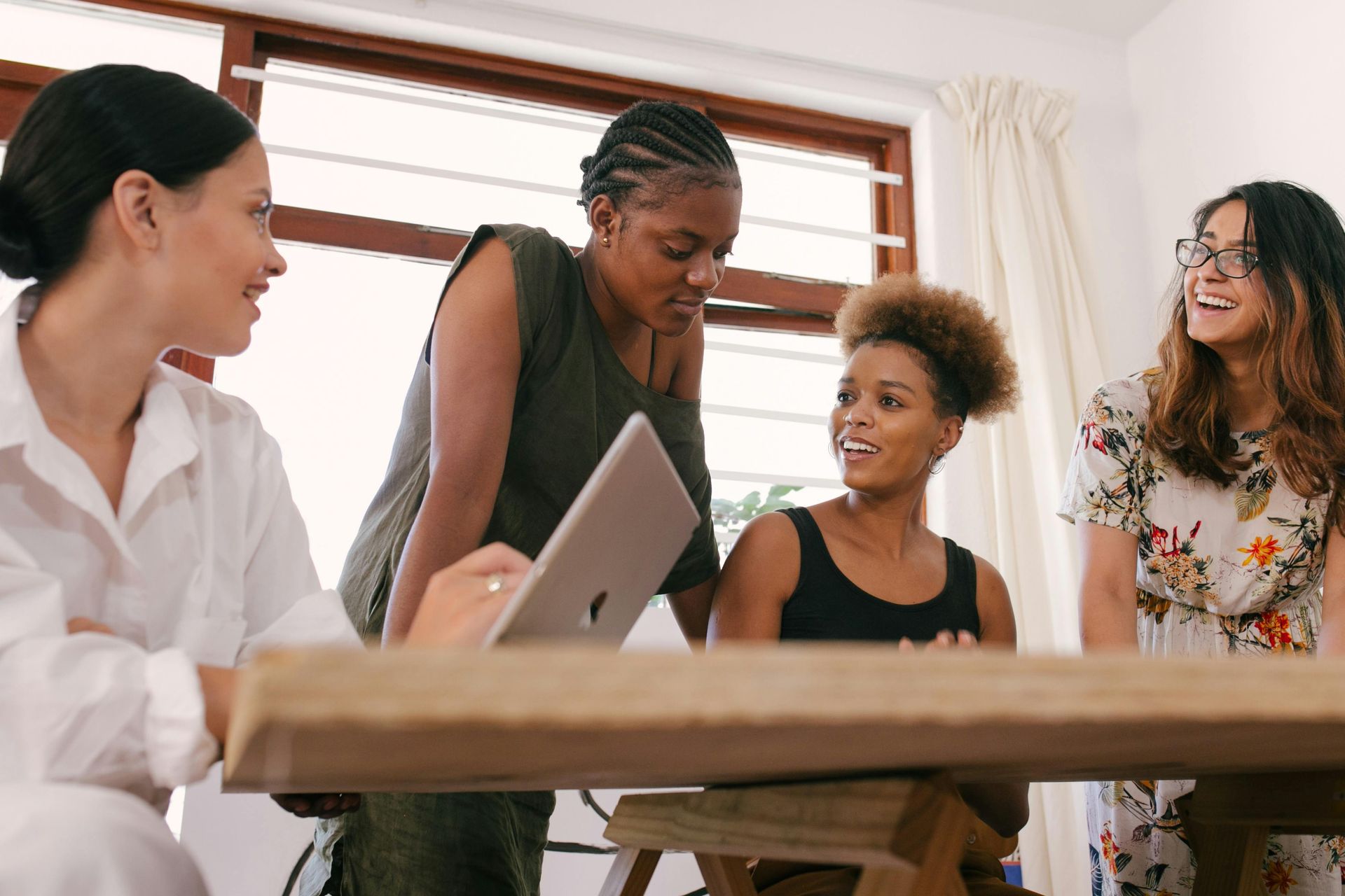 Four women gathered around a wooden table, looking at a laptop and chatting indoors.