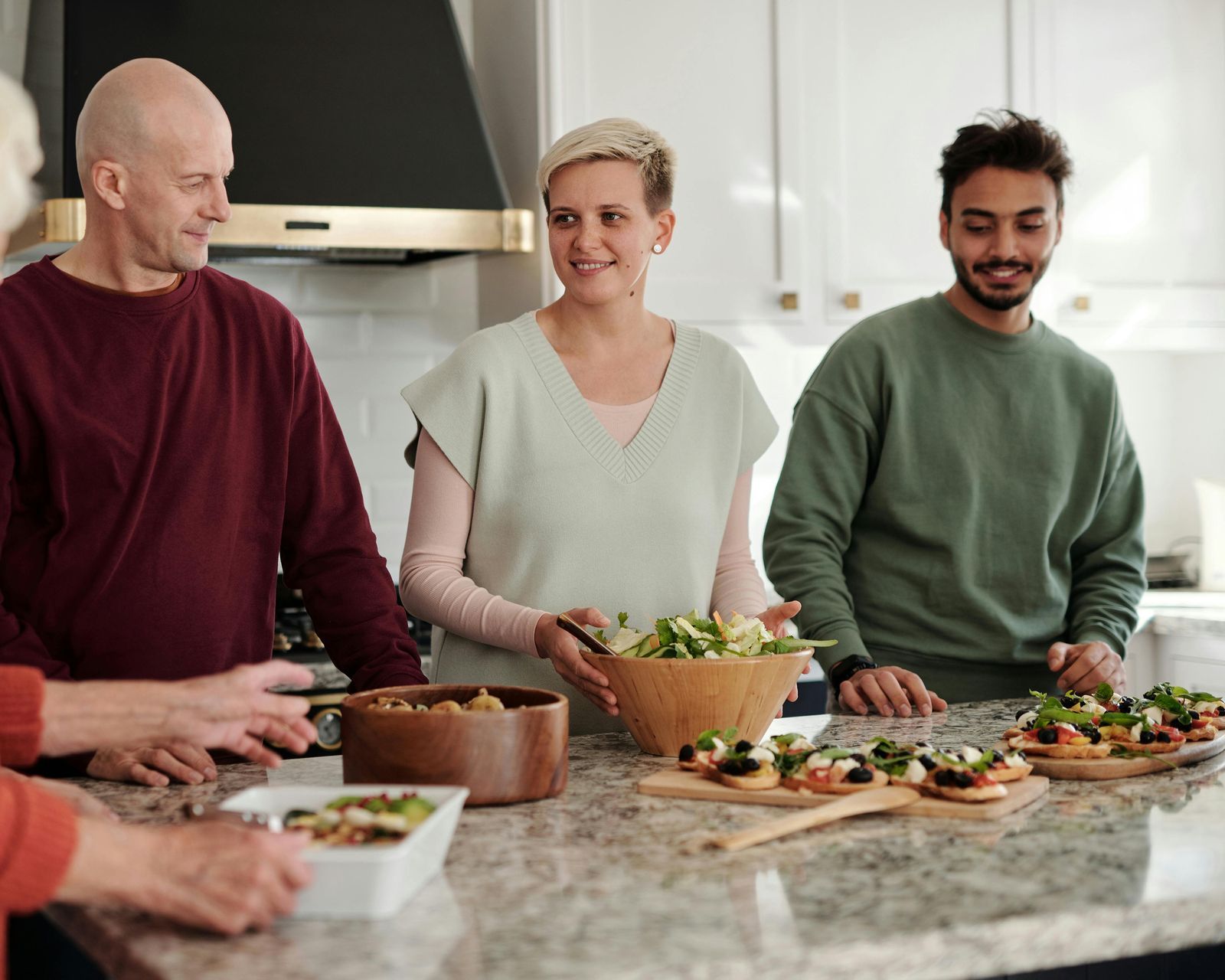 People preparing a meal in a kitchen: woman holding salad, others at countertop with food, smiling.