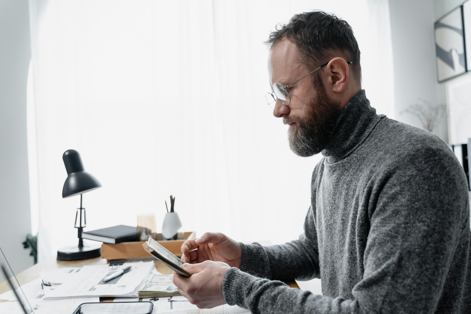 Man with beard wearing glasses and a gray turtleneck sweater looking at a tablet at a desk.
