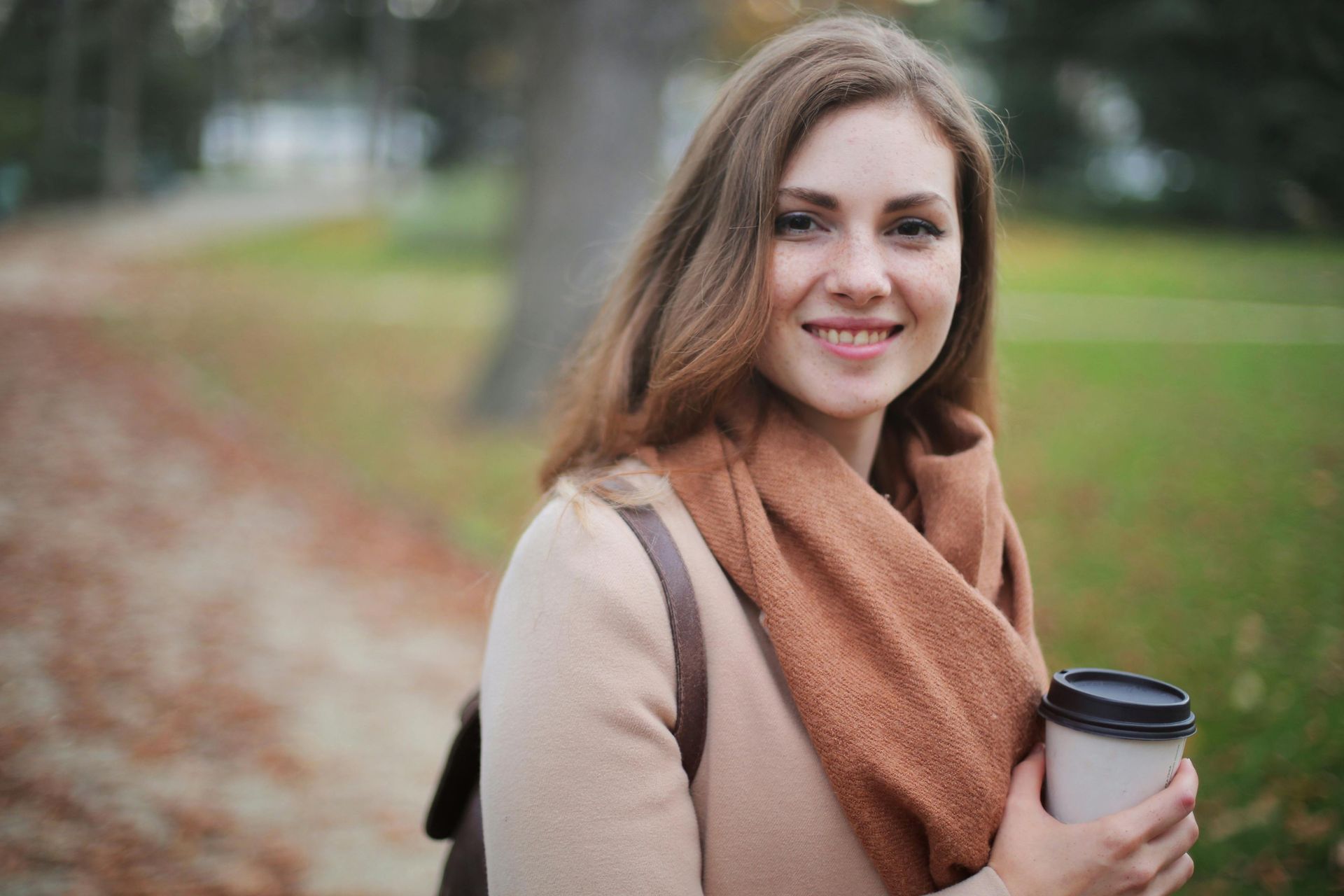 Woman smiling, holding a coffee cup, wearing a tan sweater and scarf, standing in a park with fall foliage.