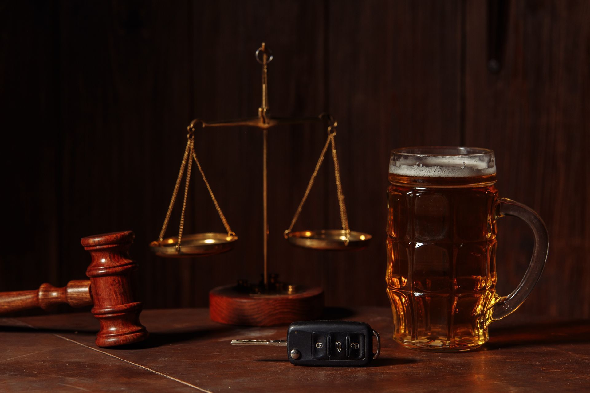 Scales of justice, gavel, car keys, and beer mug on a wooden table, suggesting DUI or legal implications of drinking and driving.