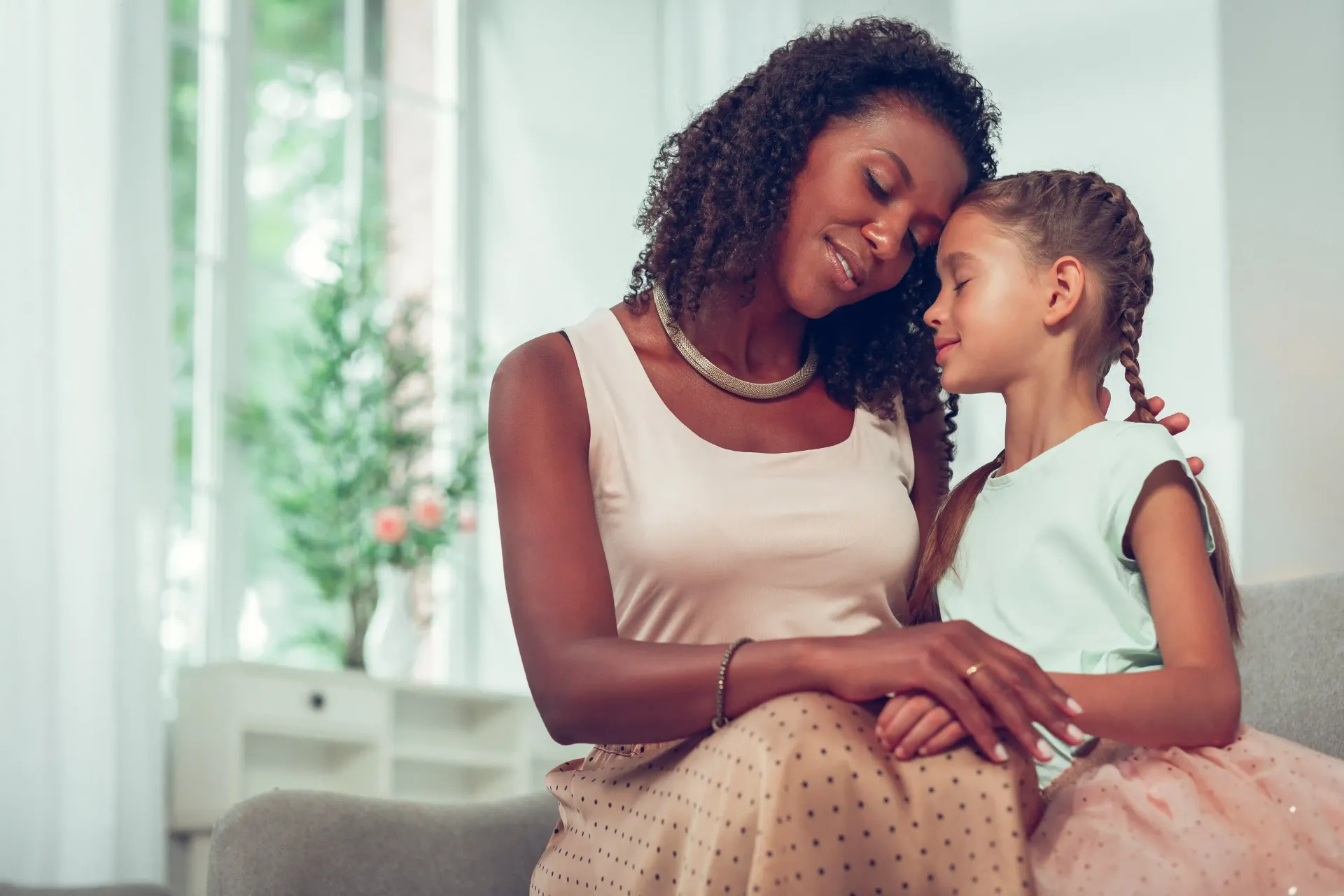 Mother and daughter embrace on a couch, both smiling; soft lighting.