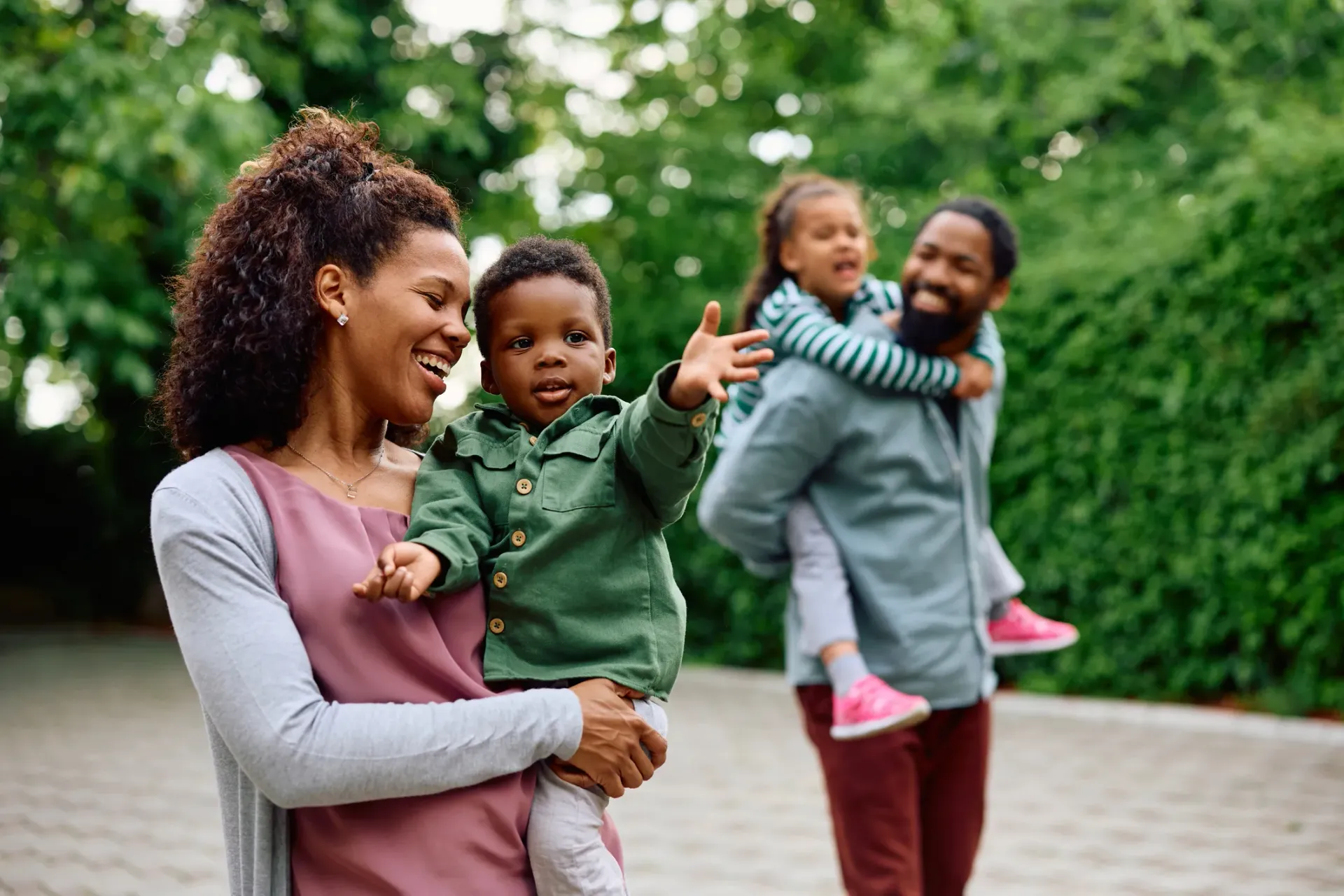 A smiling Black family enjoying the outdoors. Mom holds a toddler, waving. Dad carries another child on his back.
