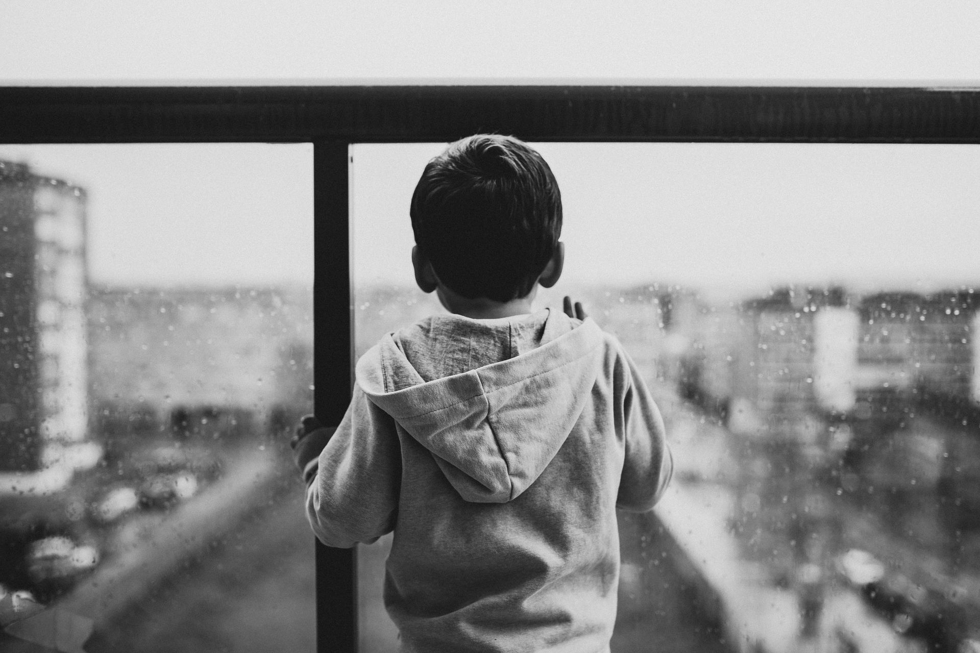 A young child in a hoodie looks out from a balcony railing towards city buildings.