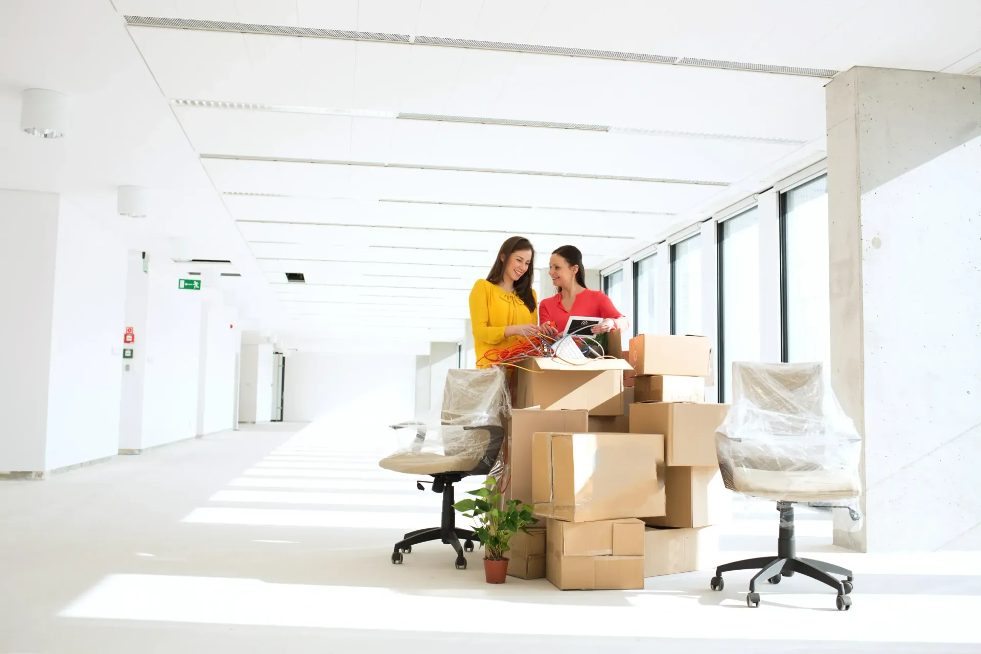 Two women are sitting at a desk in an office surrounded by cardboard boxes.