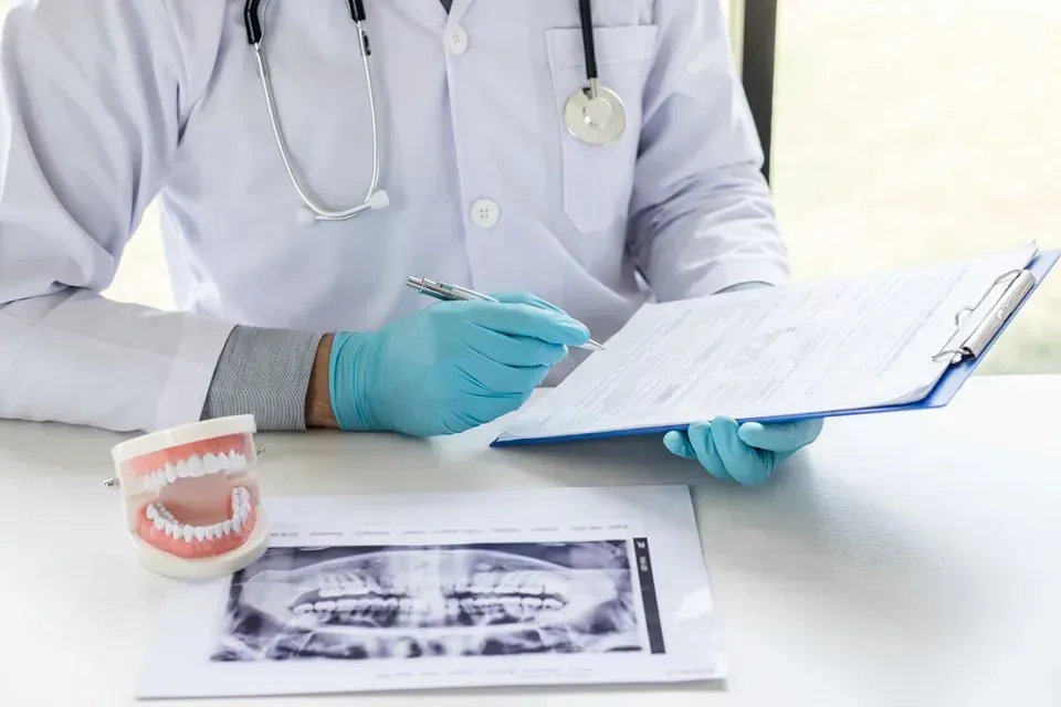 A Dentist is Sitting at a Desk With a Clipboard and a Model of Teeth — Twin Towns Denture Clinic In Gloucester, NSW