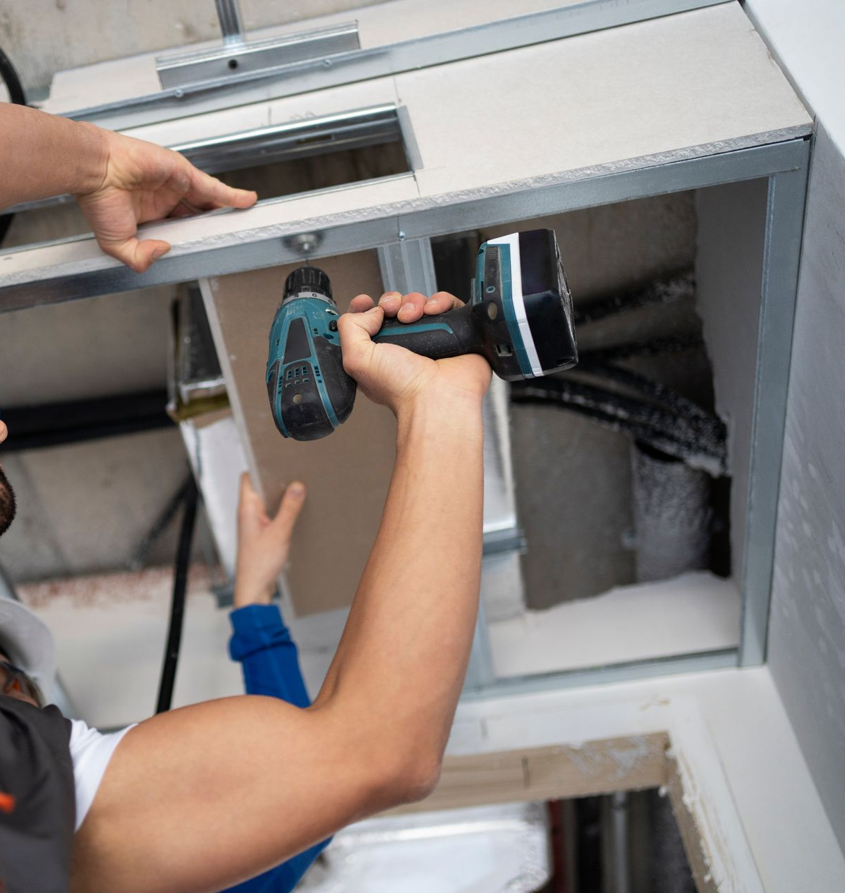 Person installing drywall with a drill on a ceiling. Another person assists.