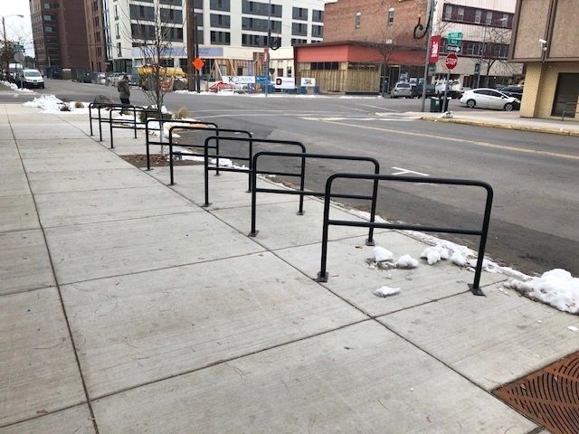 Black metal bike racks line a sidewalk next to a street with buildings in the background.