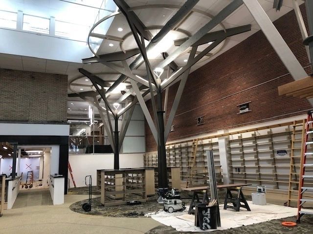 Interior of a building under construction, featuring tree-like support structures. Brick wall and work tables are visible.