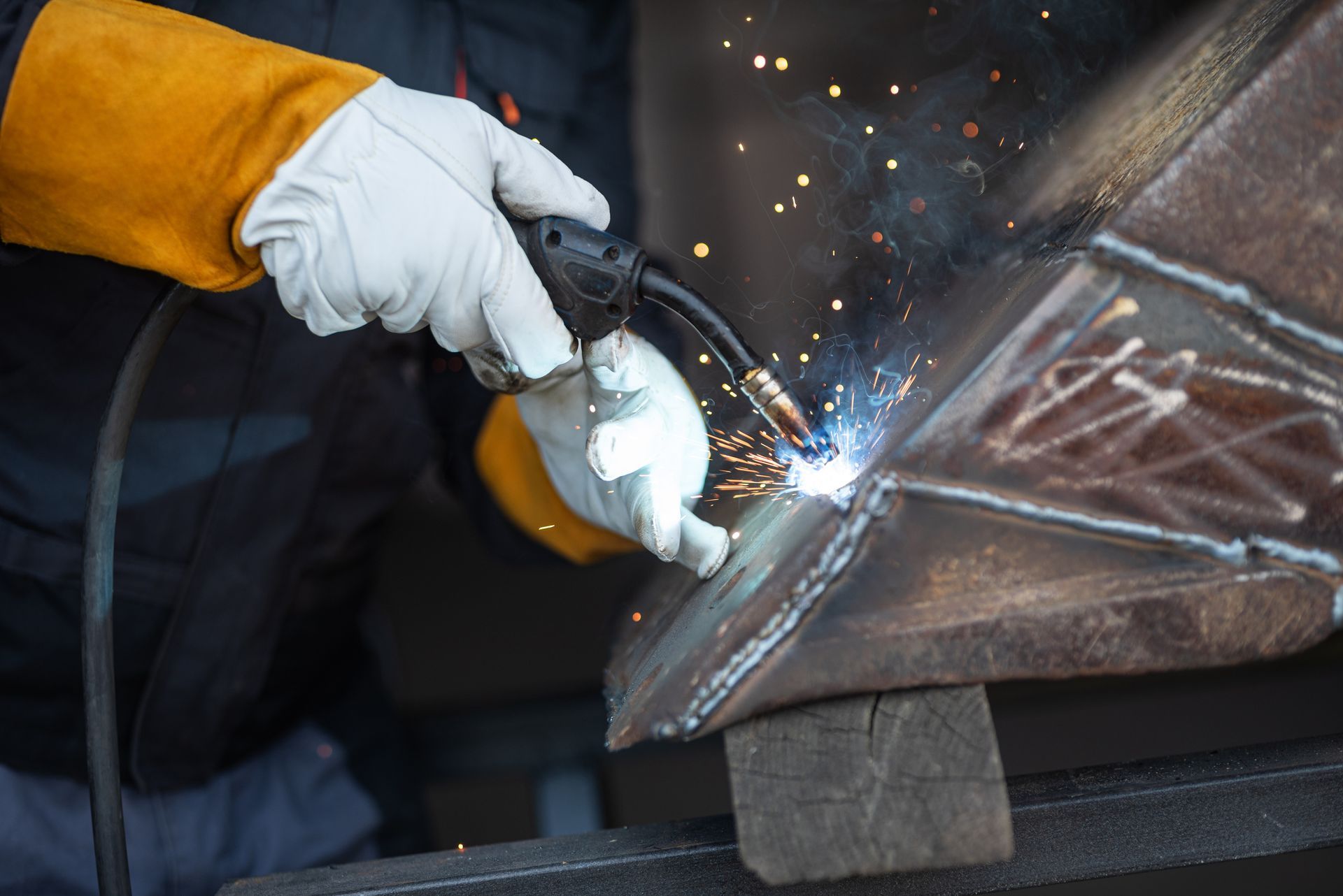 Welder in gloves using a torch, sparks flying, welding metal on a wooden support.