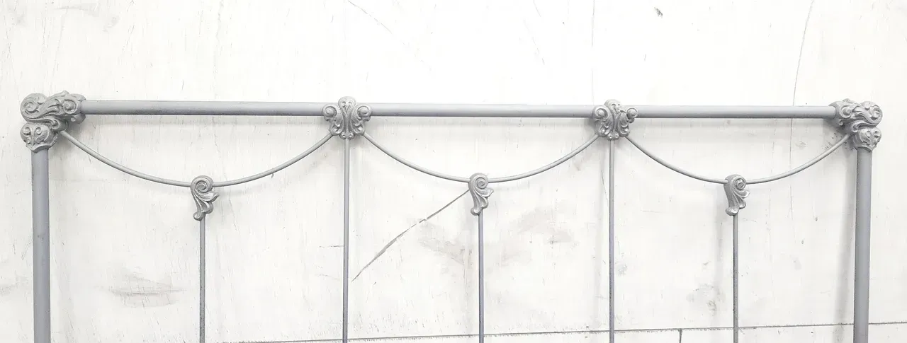 Metal bed headboard with decorative floral accents and chain details against a white wall.