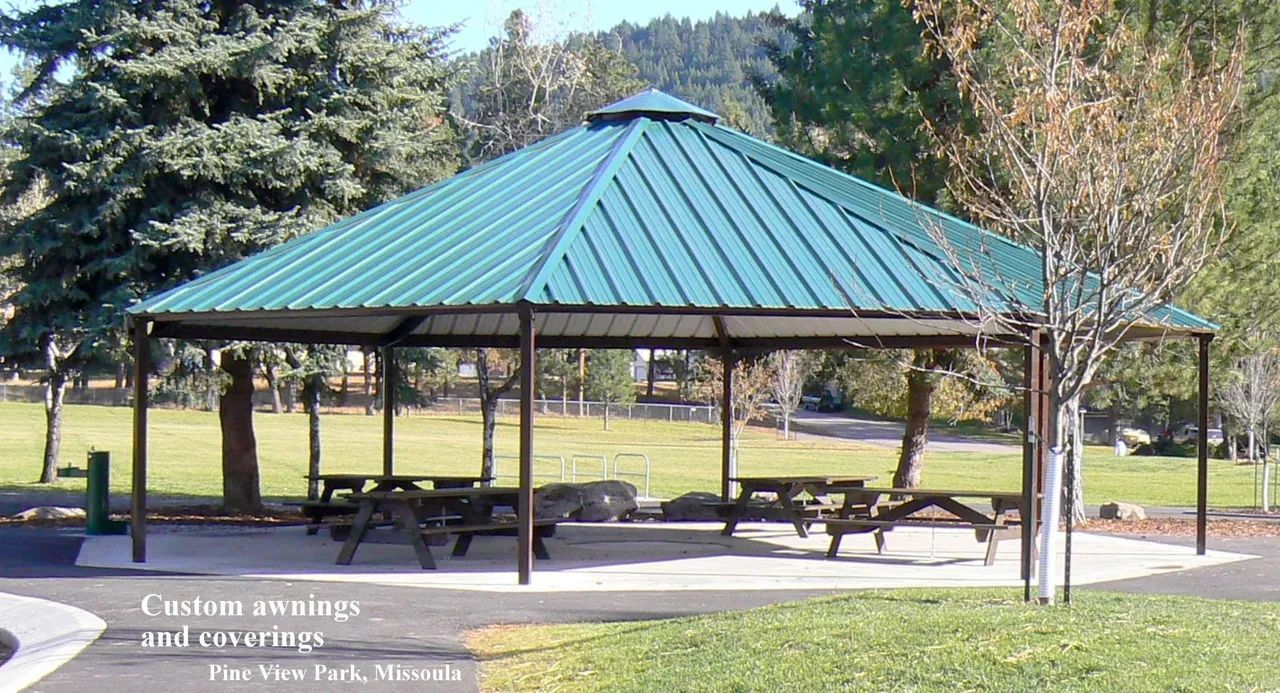 Green-roofed picnic shelter with tables on a concrete pad in a park.