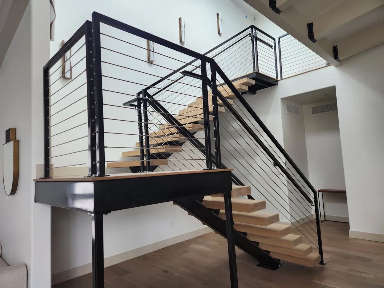 Modern staircase with wood steps, black metal railing, and cable balustrade in a white-walled interior.