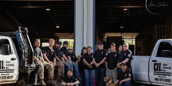 Group of people posing with two trucks in front of a garage. The people are wearing black shirts and some are smiling.